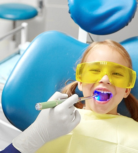 A dentist uses a curing light to harden the dental sealant placed on a young girl’s smile