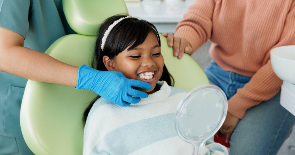 Dentist in blue gloves touching little girl's chin as she smiles in mirror